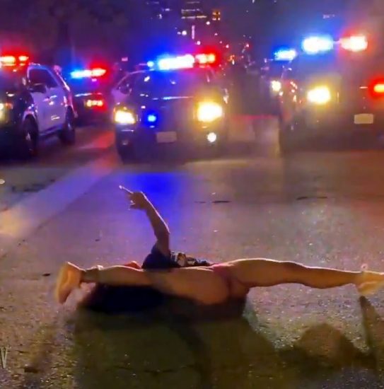 A Black woman in thong and BLM T-shirt protests by dancing and doing splits in front of a phalanx of police cars.